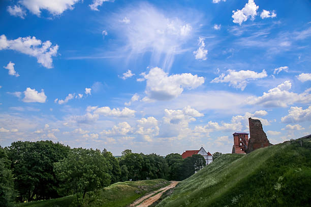 Historic ruins stand against a backdrop of bright blue sky and clouds, surrounded by lush greenery and a winding path.