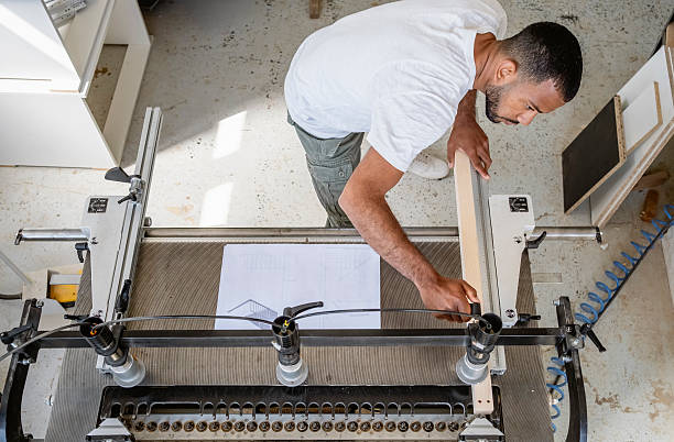 Overhead angle shows mid adult male craftsperson in white shirt operating professional wood planer machinery in sunlit workshop environment, demonstrating precise woodworking techniques.