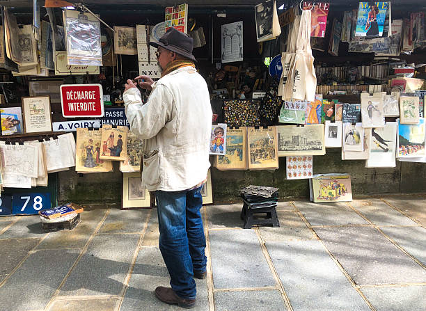 Paris, France: A sidewalk bookseller (bouquiniste) stands at his stall, surrounded by his retail display of vintage magazines, books, prints and street signs. The Seine River runs nearby, off-camera.