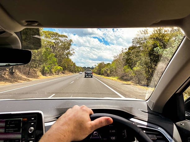 istock A wide-angle view through a car’s windscreen reveals a highway through a forested area, framed by the interior, steering wheel, and driver’s hands under a clear blue sky with fluffy clouds 2217887608