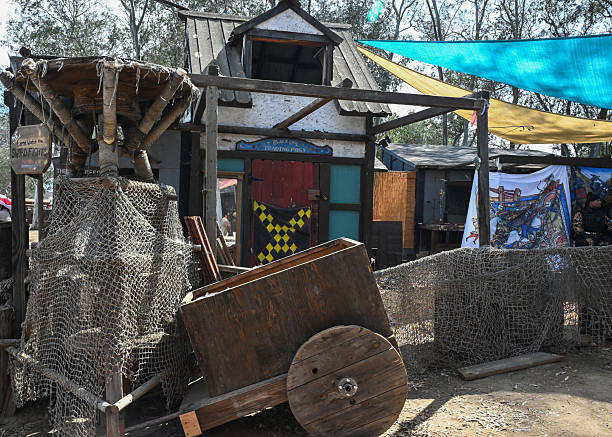 A rustic medieval trading post features a colorful storefront, fishing nets, and a wooden cart under vibrant festival canopies at a Renaissance Festival. The scene captures the atmosphere of historical reenactment, traditional craftsmanship, and the inventive design of themed festival environments.