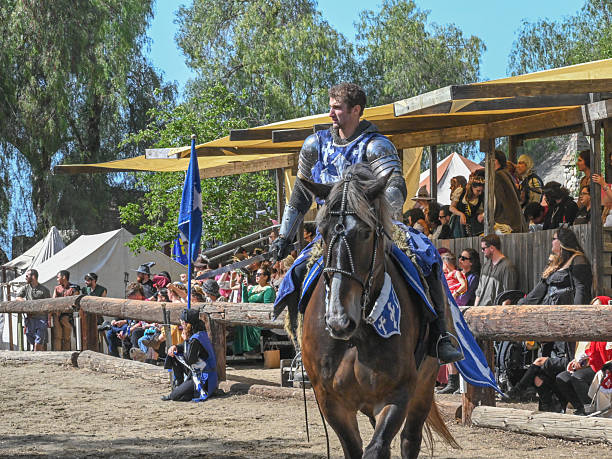 A costumed knight on horseback passes a crowd of spectators during a jousting demonstration at a renaissance fair in Eastvale, California, on May 25, 2025. The audience, dressed in both modern and period attire, watches from shaded wooden bleachers as the rider performs in full armor under bright midday sun.