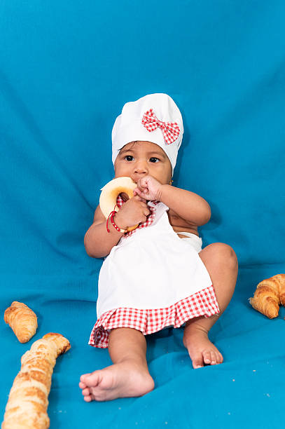 Adorable baby wearing a chef's hat and apron, enjoying a donut, surrounded by pastries on a vibrant blue backdrop