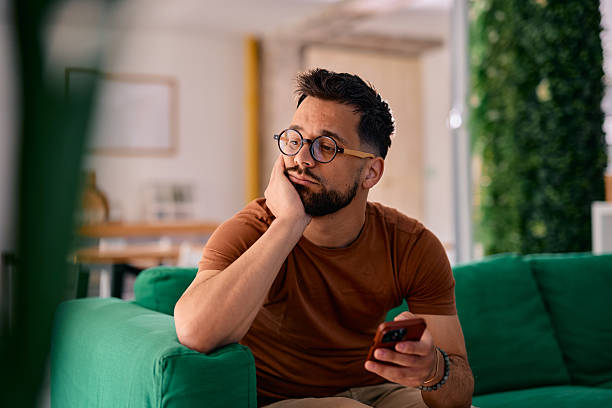 istock Pensive young man holding smartphone and looking away while sitting on sofa at home 2217946339
