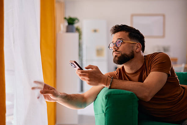 istock Pensive young man using smartphone and looking out the window at home 2217946543