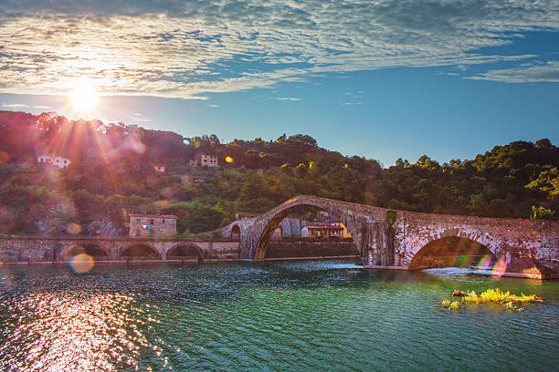 The legendary Ponte della Maddalena, known as the Devil's Bridge, creates a stunning silhouette with its distinctive asymmetrical arches spanning the Serchio River near Barga. This medieval masterpiece, steeped in local folklore and legends, glows magnificently in the warm evening light as the sun sets behind the Tuscan hills.