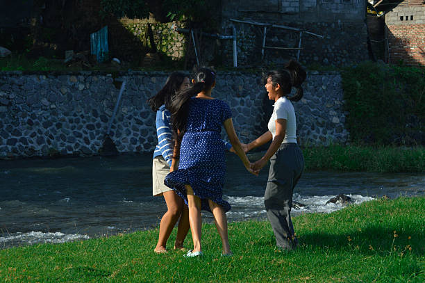 In high spirits, the girls spin around in a circle while jumping and laughing near the riverbank.