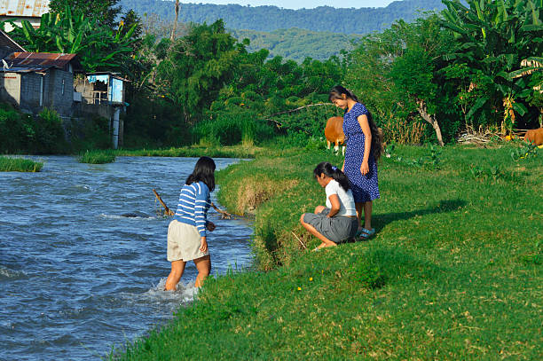 A girl squats at the waterG??s edge while her friends observe with smiles near the village stream.