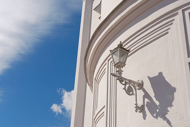 Classic street lamp casting shadow on curved white building wall. Intricate lantern attached to church or cathedral on blue sky background