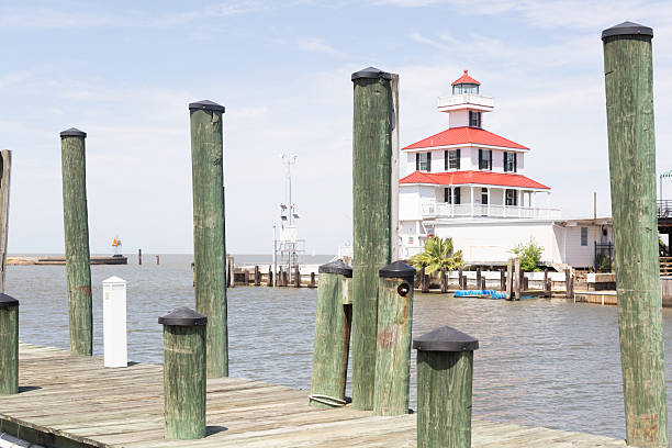 The Choptank River Lighthouse, situated at the end of Pier A at Long Wharf Park in Cambridge, Maryland, casts a reflection on the glassy waters of the Choptank River in the early morning. The white hexagon-shaped structure stands on pilings, its red roof making it easy to identify.