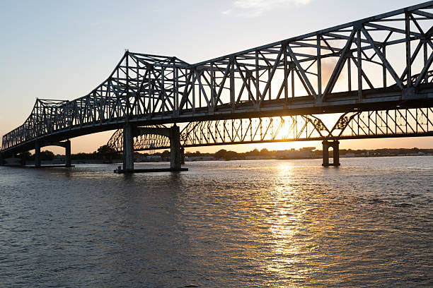 A silhouette of a large steel truss bridge spans the Mississippi River as the sun sets in the background, casting a golden reflection on the water.