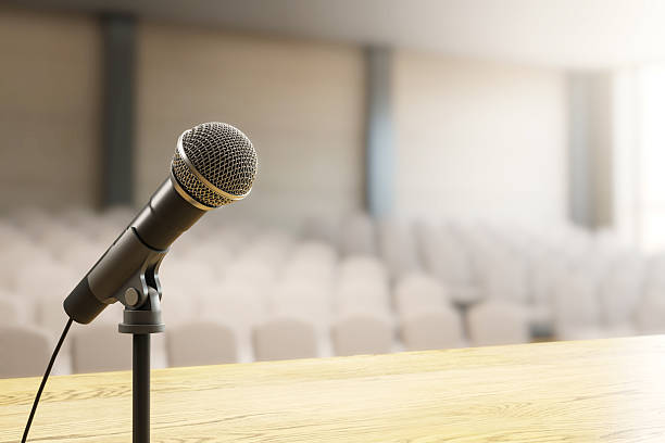 istock Microphone on wooden podium in empty conference room with blurred audience seating and soft natural light in background. 3D Rendering. 3D Rendering 2217957895