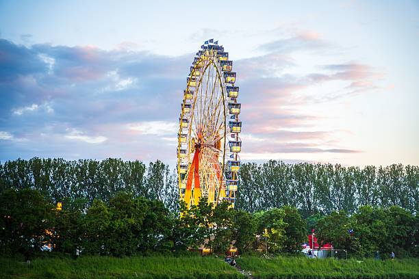 A vibrant Ferris wheel at sunset with colorful cabins against a backdrop of trees and a cloudy sky.