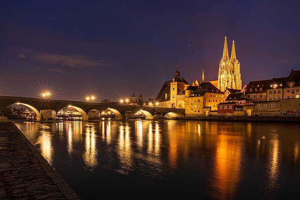 Night view of Regensburg's illuminated Stone Bridge and Cathedral reflecting in the Danube River, Germany.
