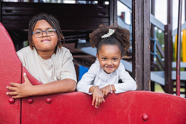 Two siblings, a boy and a girl, are smiling and leaning on a red playground fence.