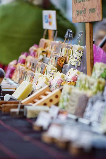Rows of colorful, decorative handmade soaps wrapped in patterned paper at a local craft fair. Wooden displays and pricing signs emphasize the artisanal and eco-friendly nature of the products.