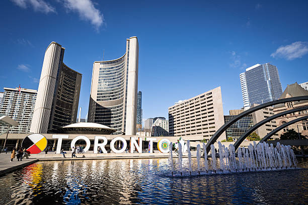 Toronto, Ontario, Canada – October 2014: People walk around the illuminated Toronto sign and fountain in Nathan Phillips Square, with the curved towers of Toronto City Hall in the background on a sunny autumn day.