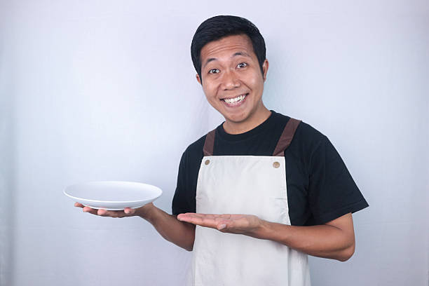 istock Smiling asian man chef or waiter shows empty white plate, presenting something 2217977141