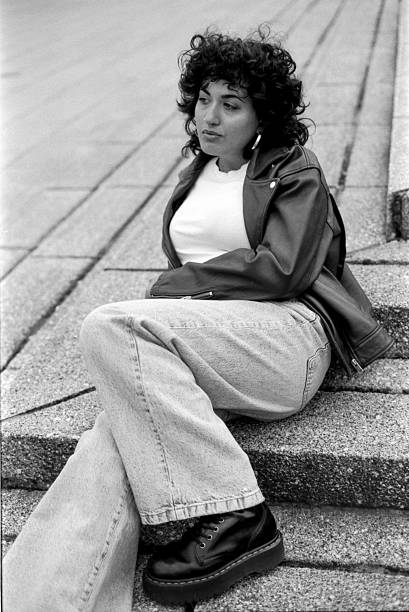 Grainy monochrome analog vintage photography from the 90s taken with 35mm film camera, portraying a young woman wearing a leather jacket and jeans sitting on concrete steps