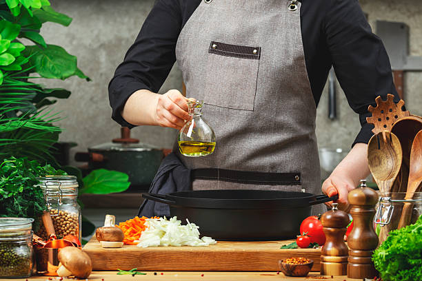 istock Chef holds olive oil jar over saucepan. Cozy kitchen with wooden table, kitchenware, vegetables, herbs and ingredients for cooking. Healthy food, eating, culinary, recipes, food blogging 2217987284