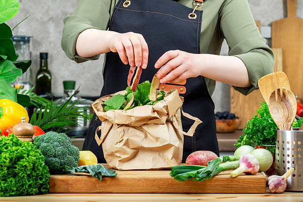 istock Chef puts vegetables peels in paper package for processing compost. Wooden table, kitchenware, vegetables and herbs. Healthy eating, zero waste, blogging, recycling 2217987305