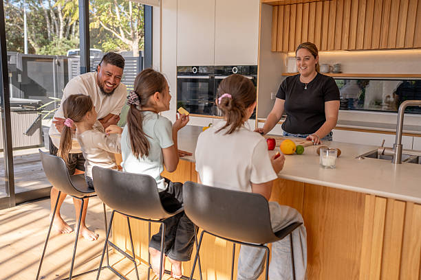 A cheerful family gathers in a modern kitchen, with three children sitting at the island enjoying fruit while chatting with their parents.