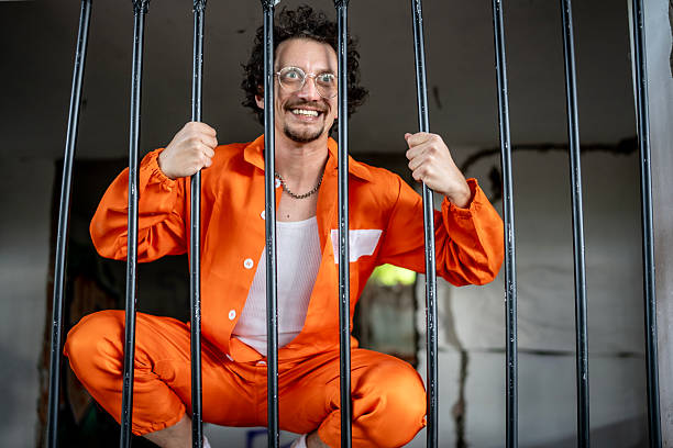 istock Cheerful Prisoner in Orange Jumpsuit Posing Behind Jail Cell Bars 2218009755