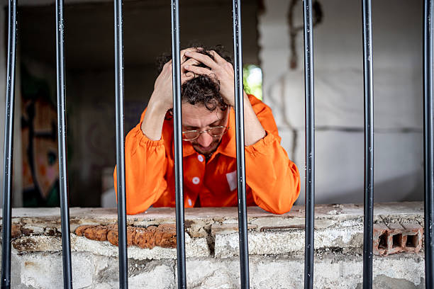 istock Man in Prison Cell Appearing Distressed, Wearing Orange Jumpsuit Behind Bars 2218009760