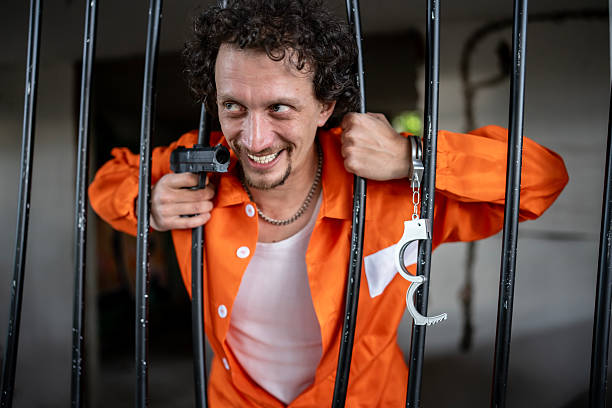 A man wearing an orange prison jumpsuit leans against jail cell bars, holding a gun and handcuffs. The expression and setting suggest dramatization of criminal or escape scenarios in a confined environment.