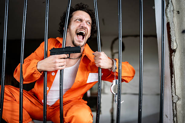 istock Prisoner Holding Handgun Behind Bars in Orange Jumpsuit 2218009779