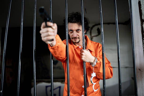 istock Man in Prison Uniform Pointing Gun Behind Bars in Confinement Cell 2218009789