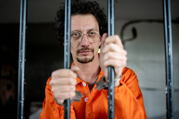 istock Man in Orange Prison Jumpsuit Behind Bars in Dimly Lit Setting 2218010051