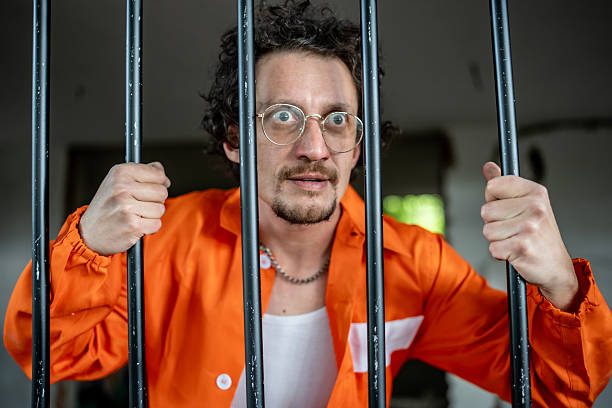 istock Man in Prison Uniform Stands Behind Bars in a Dimly Lit Room 2218010082