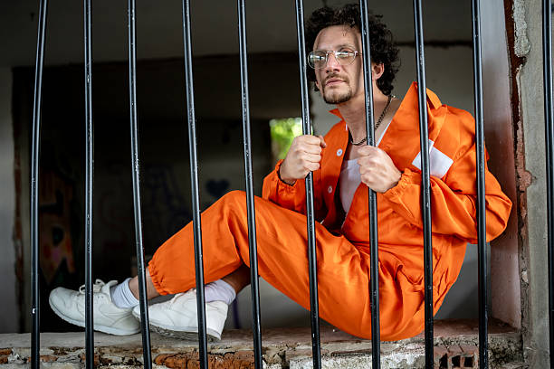 istock Man in Orange Uniform Sitting Behind Prison Bars Looking Thoughtful 2218010083