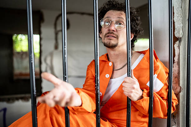istock Man in Prison Cell with Orange Jumpsuit Looking Through Bars 2218010101