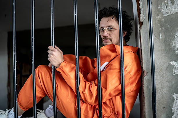 A reflective individual in an orange jumpsuit is seated against bars in a deteriorated prison setting, capturing themes of confinement, solitude, and introspection.
