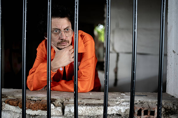 istock Man Wearing Orange Jumpsuit Looking Through Bars in a Prison Setting 2218010122