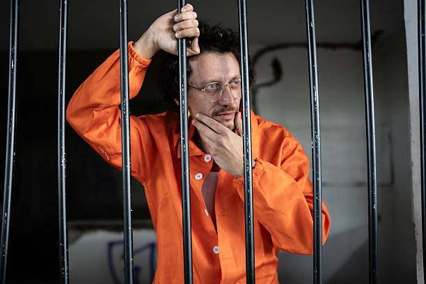istock Man in Orange Uniform Standing Behind Bars in Prison Cell 2218010127