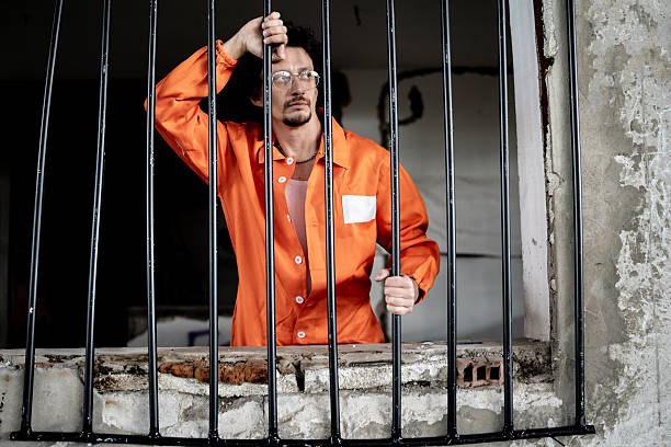 istock Man in Orange Jumpsuit Standing Behind Prison Bars in a Concrete Environment 2218010136