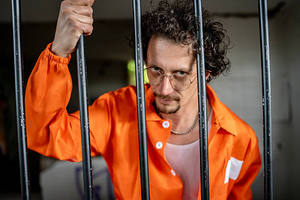 istock Prisoner in Orange Jumpsuit Standing Behind Bars in a Jail Cell 2218010149