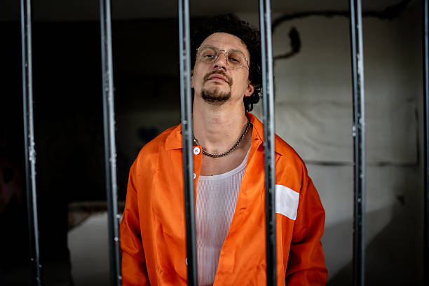 istock Man Wearing Orange Prison Uniform Standing Behind Jail Bars 2218010152