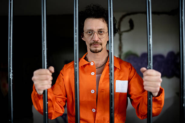 istock Man in Orange Prison Uniform Stands Behind Jail Bars 2218010153