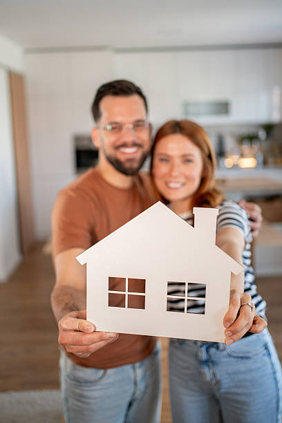 istock Happy couple holding a small white house model, smiling at the camera 2218016495