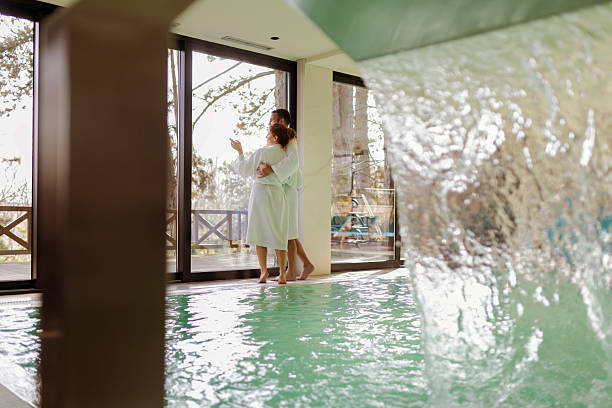 A Couple Stands Together in a Modern Spa Center, Embracing as They Take in the Beauty of Nature Through a Large Glass Window. A Representation of Love, Relaxation, and Wellness.