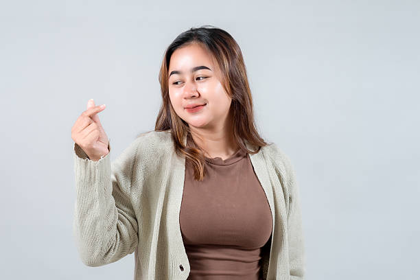 istock A young woman smiles brightly while making a playful hand gesture, often associated with love or affection. She wears a cozy sweater, exuding a cheerful and friendly vibe against a neutral background 2218071772