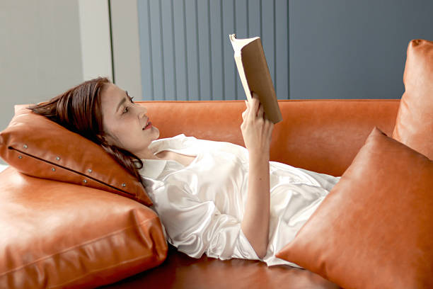 Happy middle aged female reading book while lying down on sofa in living room. Beautiful relaxing adult Asian woman spends her free time reading novel book at home. Smiling woman enjoy reading book.