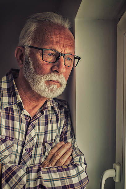 Portrait of the depressed and sad senior man standing beside window.