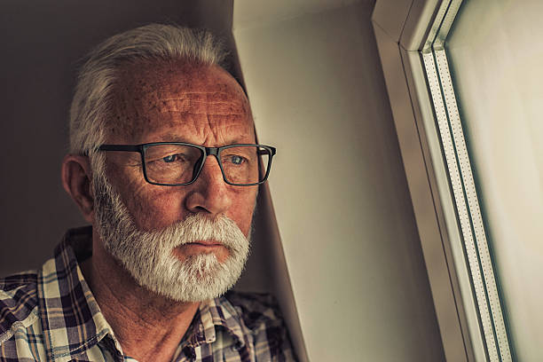 Portrait of the depressed and sad senior man standing beside window.