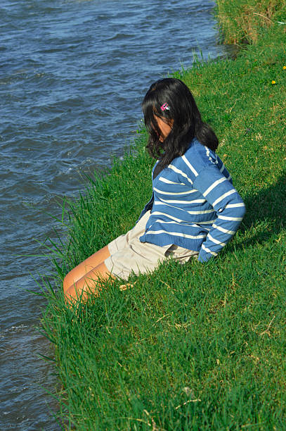 A teenage girl sits alone by the riverside, dipping her feet in the water and enjoying the quiet moment.
