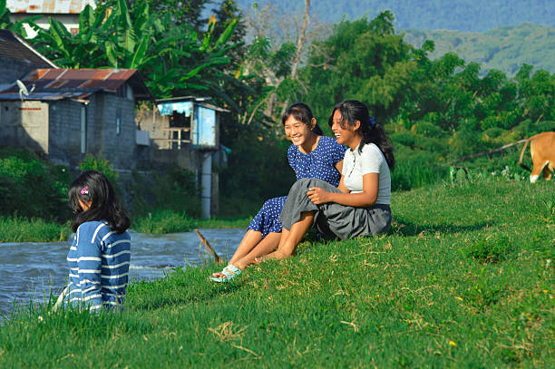 Two girls sit on the grassy field laughing while their friend plays by the waterG??s edge.